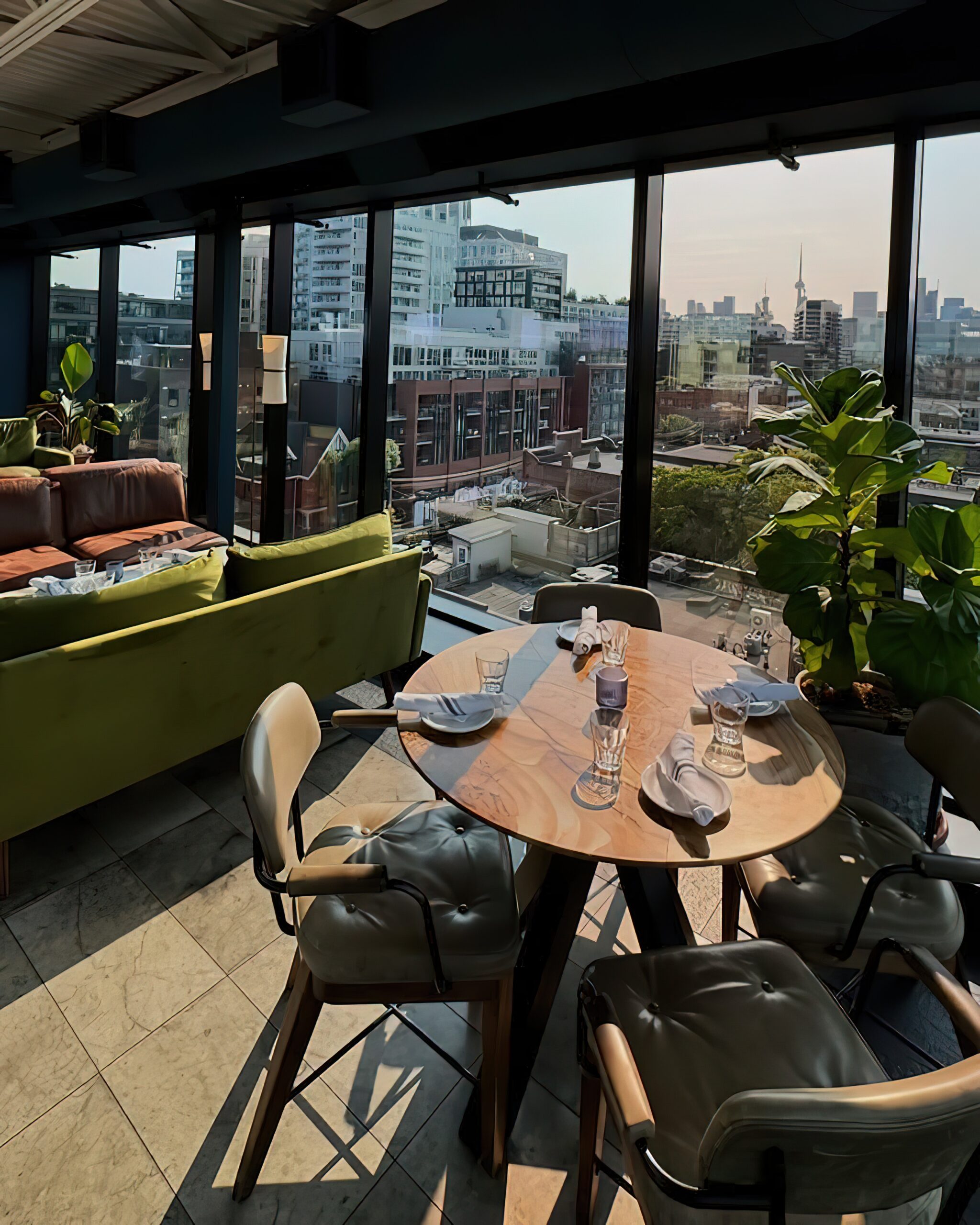 The Rooftop at the Broadview Hotel | Golden hour shot of the atrium with floor to ceiling windows, at round table set for 4 with green chairs, green low slung couches and plants | Homes Almanac