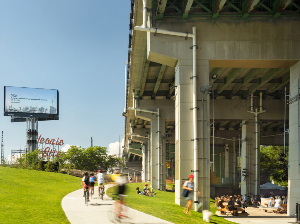 Beneath the Gardiner at The Bentway | Bright sunny day with people biking, walking and lounging under the bentway next to a grassy hill | Homes Almanac