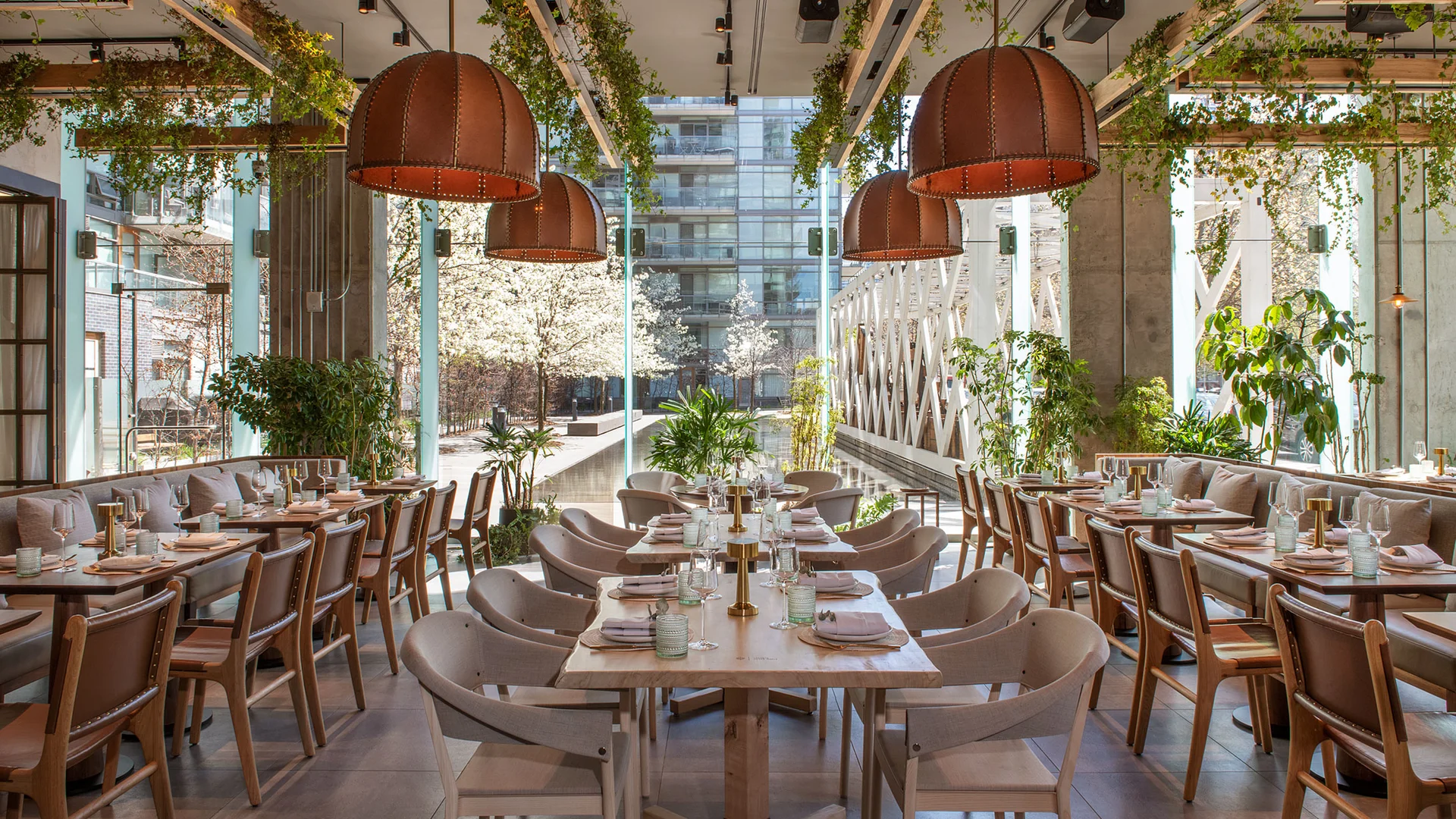 Casa Madedra | Daytime shot of the restaurant with long tables and chairs in a light taupe colour against floor to ceiling windows and leafy green plants hanging from the ceiling | Homes Almanac
