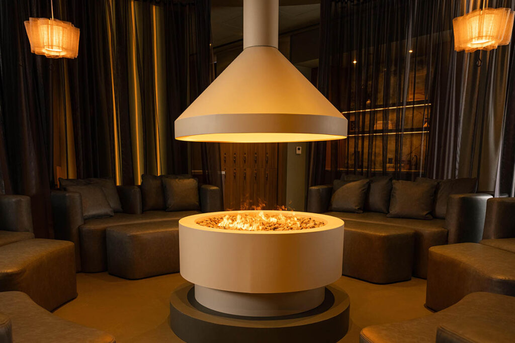 trove wellbeing | a white circular fireplace with a small fire going surrounded by low green leather sofas and ottomans in a dimly lit room | Homes Almanac