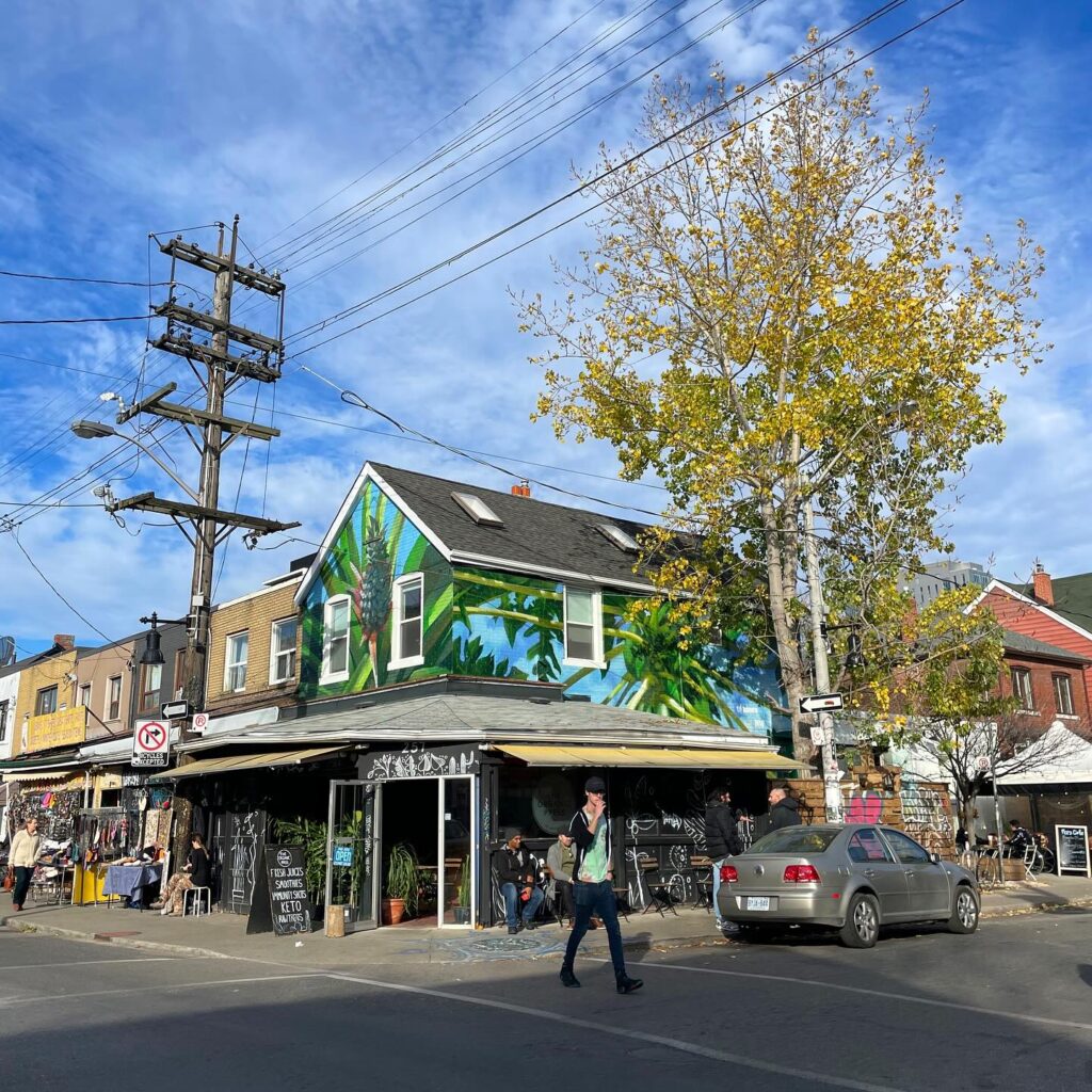 Layers of Kensington Market | Blue sky with a building with a blue and green mural painted on it and a mature tree next to the building | Homes Almanac