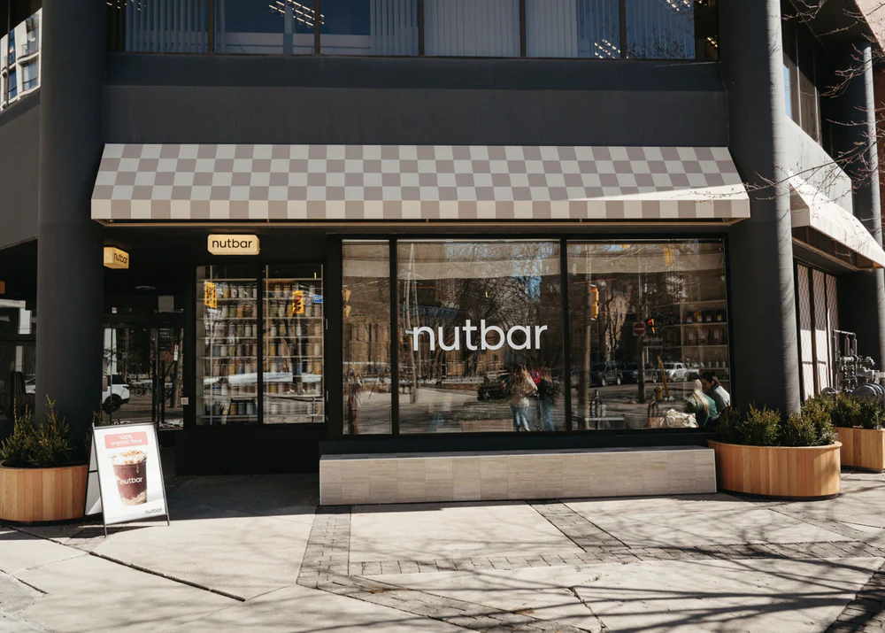 Nutbar on Adelaide | Sunny exterior shot of the shop with floor to ceiling windows and a checkered white and almond coloured awning | Homes Almanac