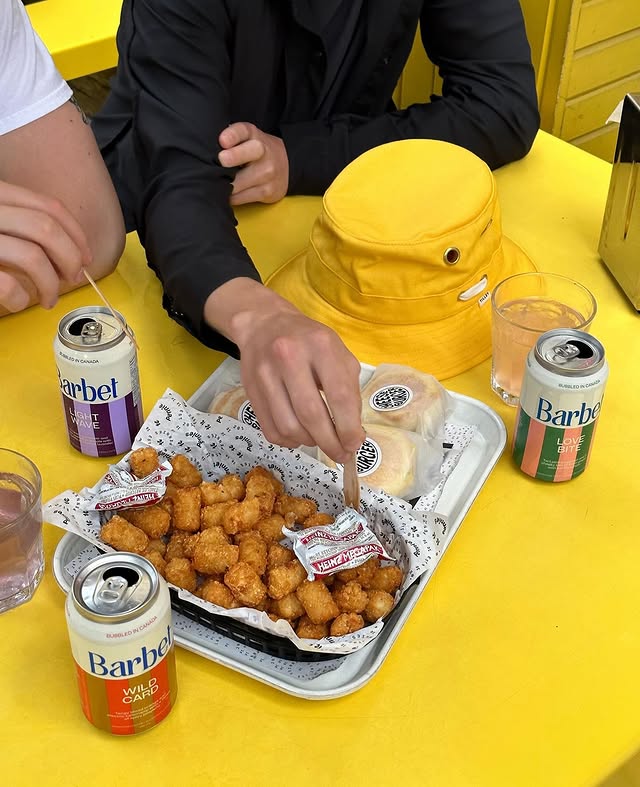 Pennies Toronto | Overhead shot of tater tots and burgers on a tray sitting on a yellow table with Barbet soda cans | Homes Almanac