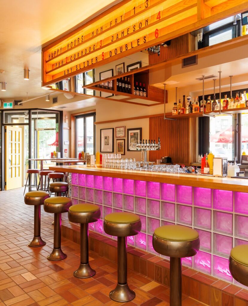 Pennies Toronto | Warmly lit bar and seating with hanging alcohol shelves, bar stools and a pink lit up bar | Homes Almanac