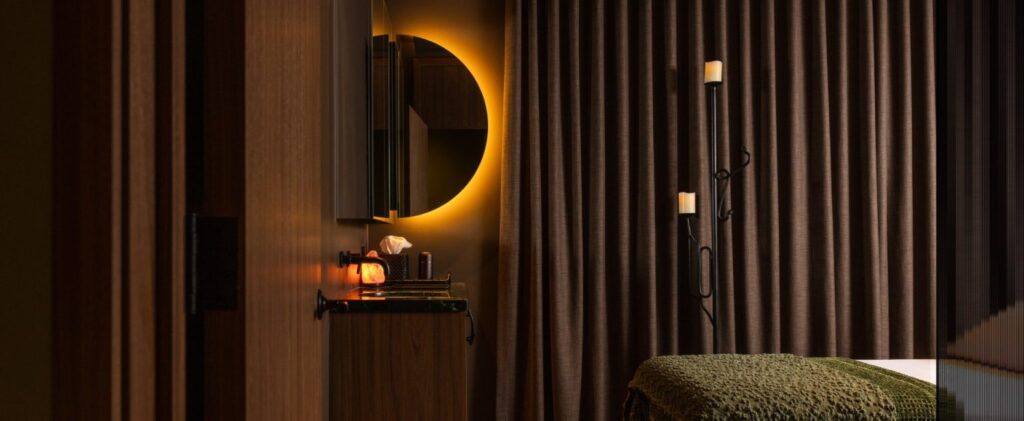 Trove Wellbeing | Dimly lit massage treatment room with brown floor to ceiling curtains, a circle mirror on the wall and a bed with green blanket to the right | Homes Almanac