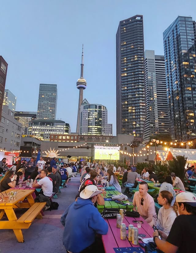 Rendeziews | Evening shot of an outdoor event venue with multicoloured picnic benches and people sitting with the CN tower in the background | Homes Almanac