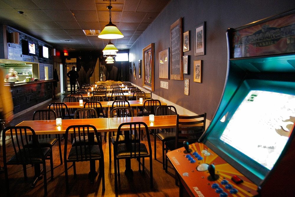The Dock Ellis | A dimly lit bar with wood tables and metal chairs and an arcade game in the corner | Homes Almanac