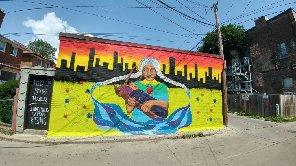 A brightly coloured mural with a yellow susnet background and a woman with 2 braids holding a baby | queen street toronto street art | Homes Almanac