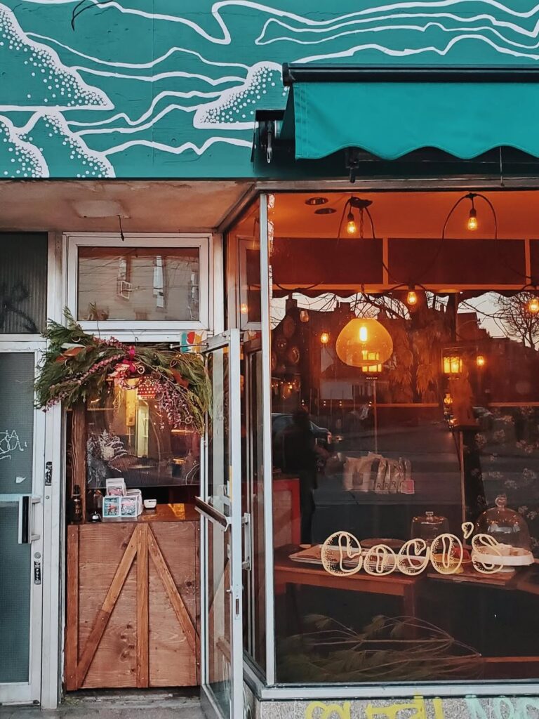 Coco's Neighbourhood Coffee Shop | Quaint exterior of a coffee shop with green awning and arm lighting through the windows | Homes Almanac