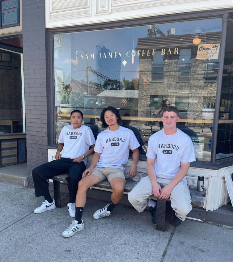 Sam James Coffee Bar on Harbord | 3 baristas in white tshirts sitting out front of the coffee shop smiling at the camera | Homes Almanac