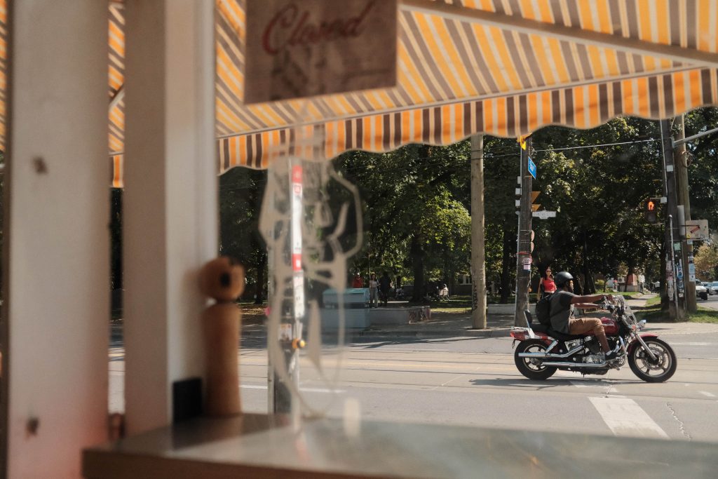 history of trinity bellwoods and ossington | through a glass window a motorcyclist moves across the intersection on a sunny day looking across to the park | homes almanac