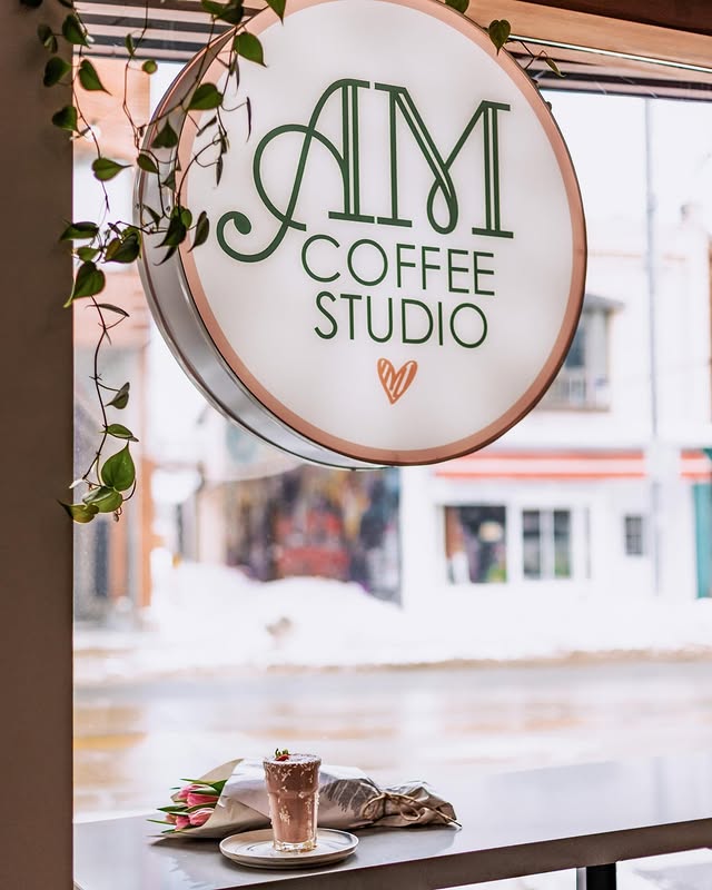 AM Coffee Studio | A circle sign with "AM Coffee Studio" written on it hangs infront of a window with a wood table and a coffee sit | Homes Almanac