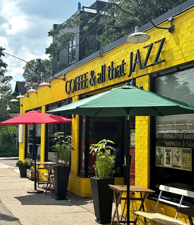 Coffee and All That Jazz | A bright yellow brick facade with green and yellow outdoor umbrellas and two planters on either side of the door on a sunny day | Homes Almanac